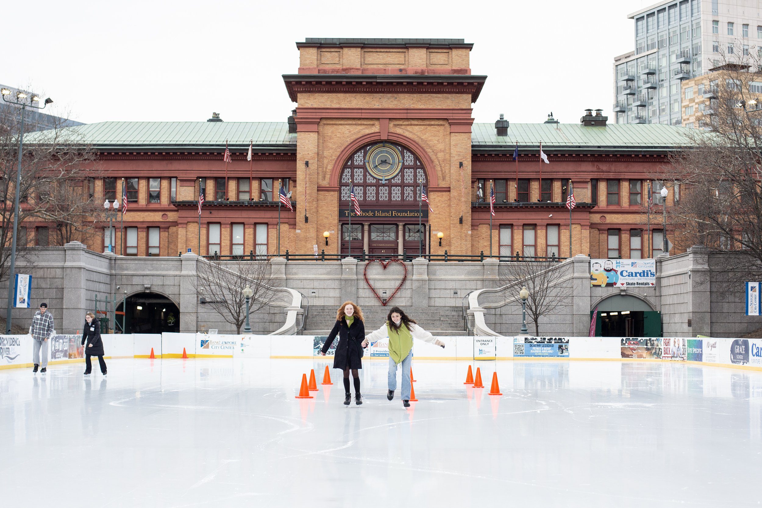 Two people ice skating at the Providence Rink in front of the historic Rhode Island Foundation building, showcasing a vibrant community winter activity with a heart-shaped decoration on the steps.